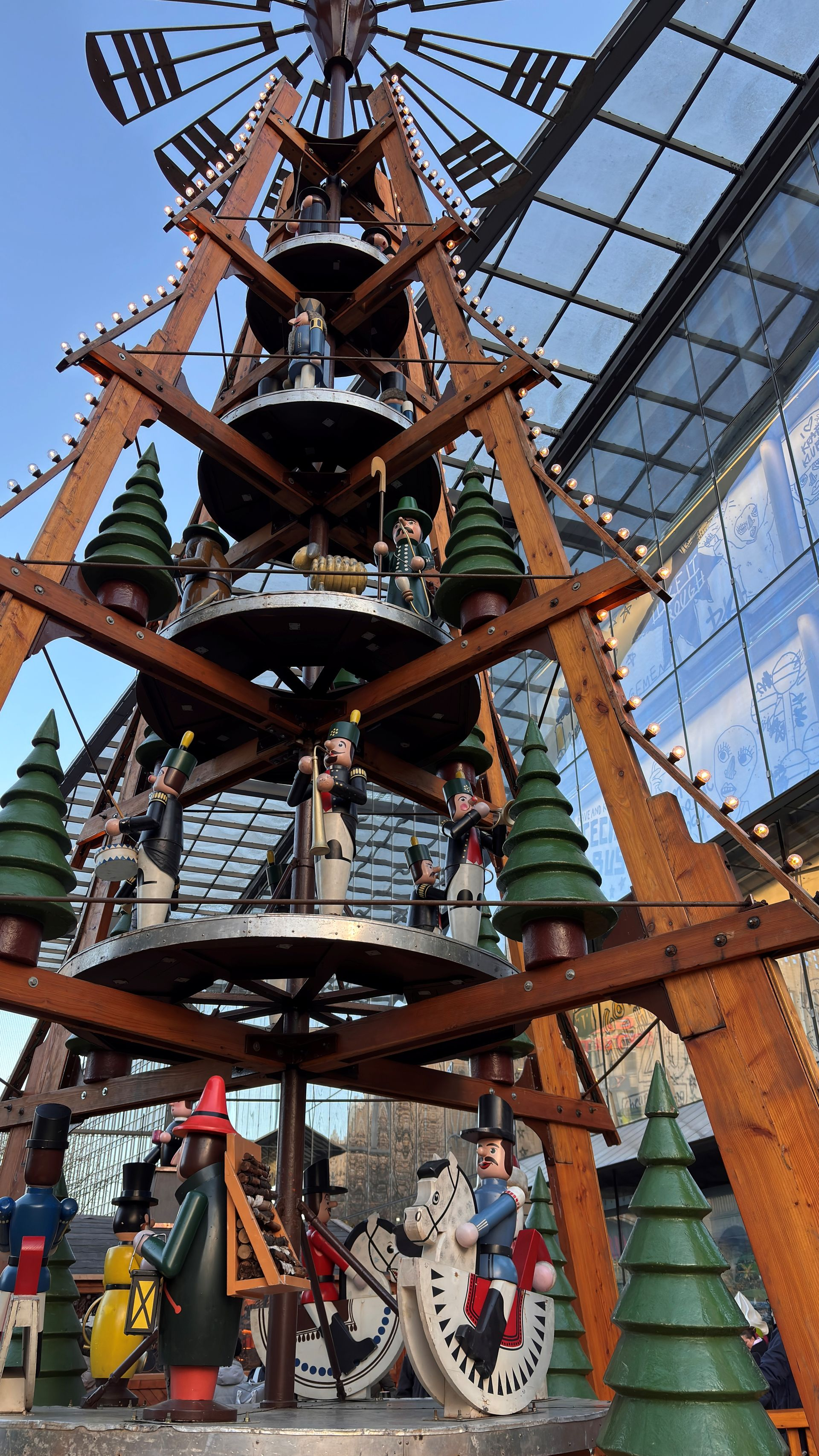 Steinturm mit Zifferblättern, gekrönt von einem dunklen Dach, eingerahmt von grünen Bäumen vor blauem Himmel.