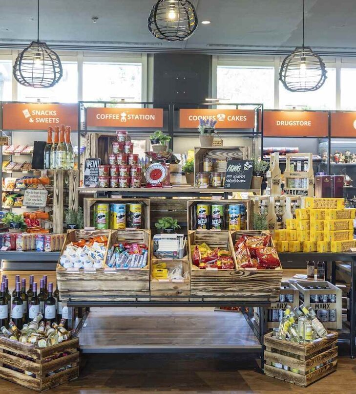 Warenauslage in einem Lebensmittelgeschäft mit Snacks, Kaffee und Getränken in Holzkisten unter Deckenbeleuchtung.