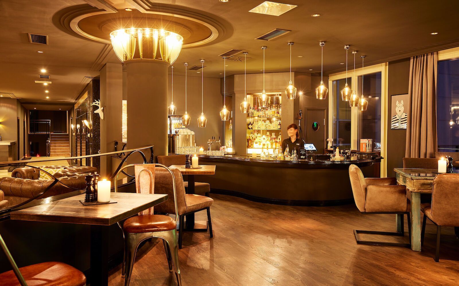 Warm beleuchtetes Bar-/Restaurantinterieur mit Gästen an Tischen, Barkeeper an der Bar, Decken- und Pendelleuchten.