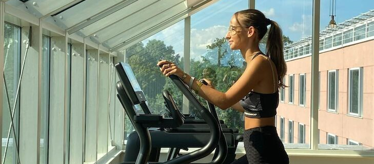Eine Person benutzt einen Ellipsentrainer in einem Fitnessstudio mit großen Fenstern und Blick auf die Bäume.