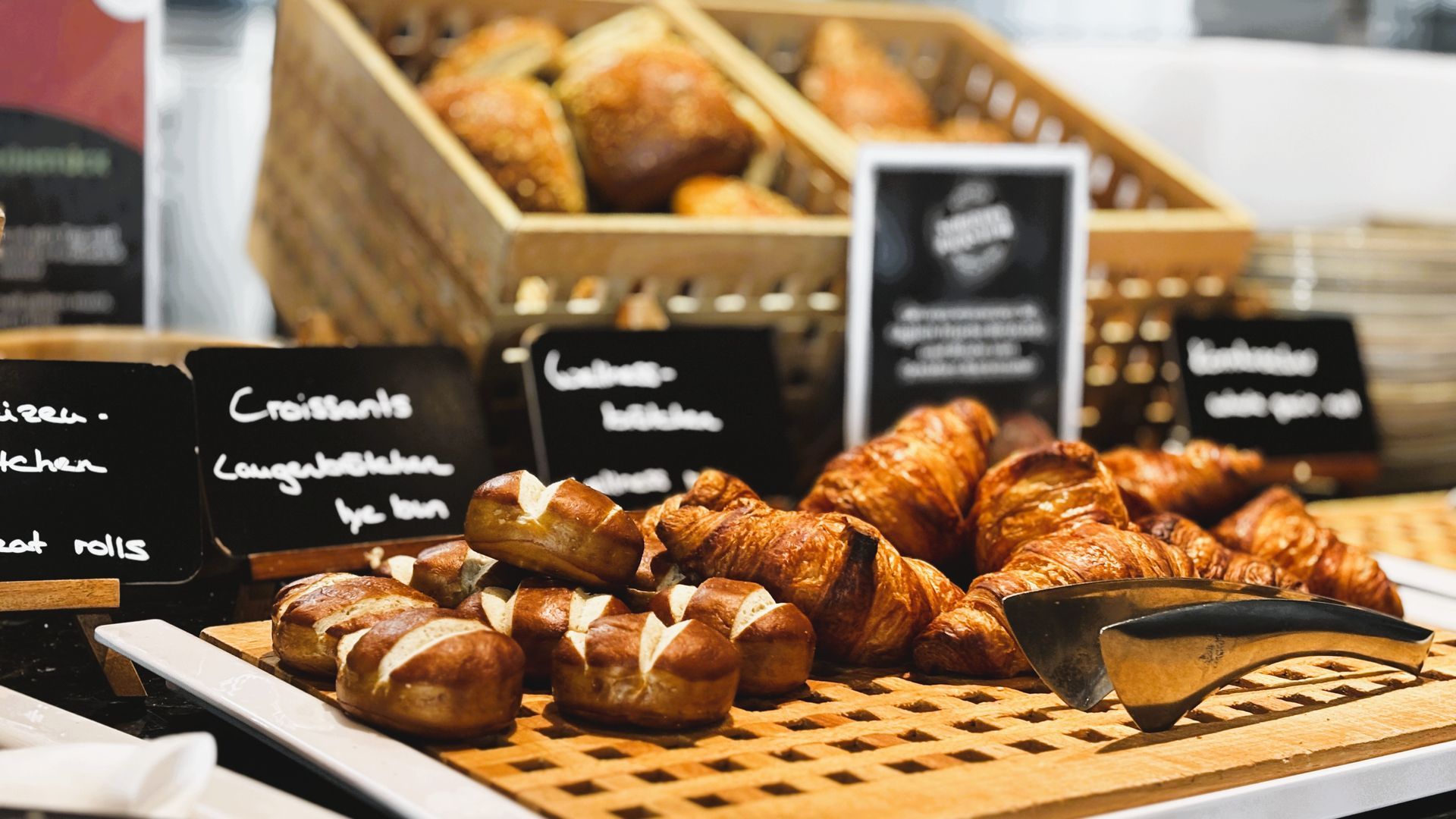 Bäckereiauslage mit Croissants, Schokoladengebäck und Brotlaiben auf Tabletts mit Kreidetafeln.