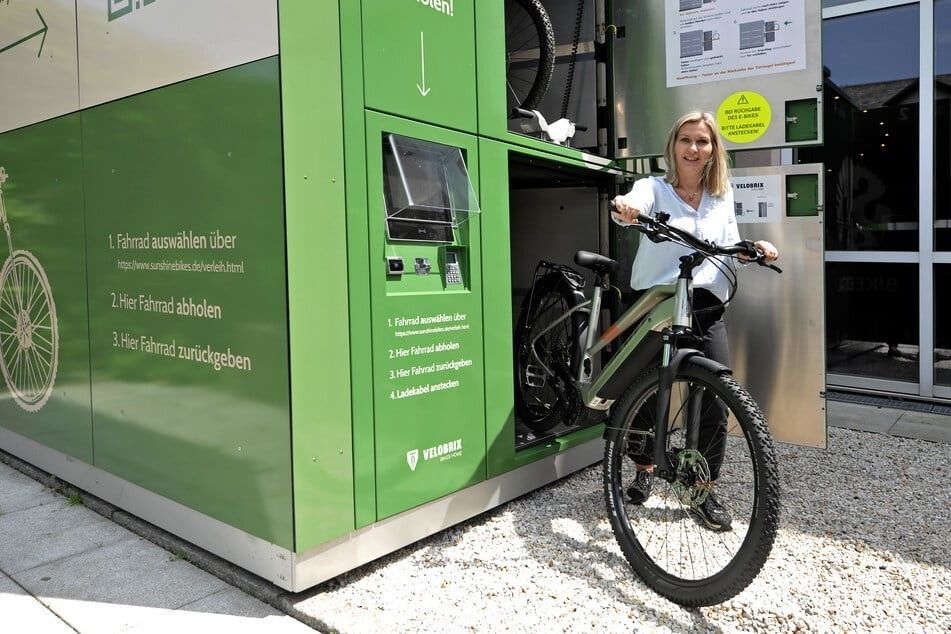 Frau mit Fahrrad an einer grünen Fahrradverleihstation vor einem Gebäude.