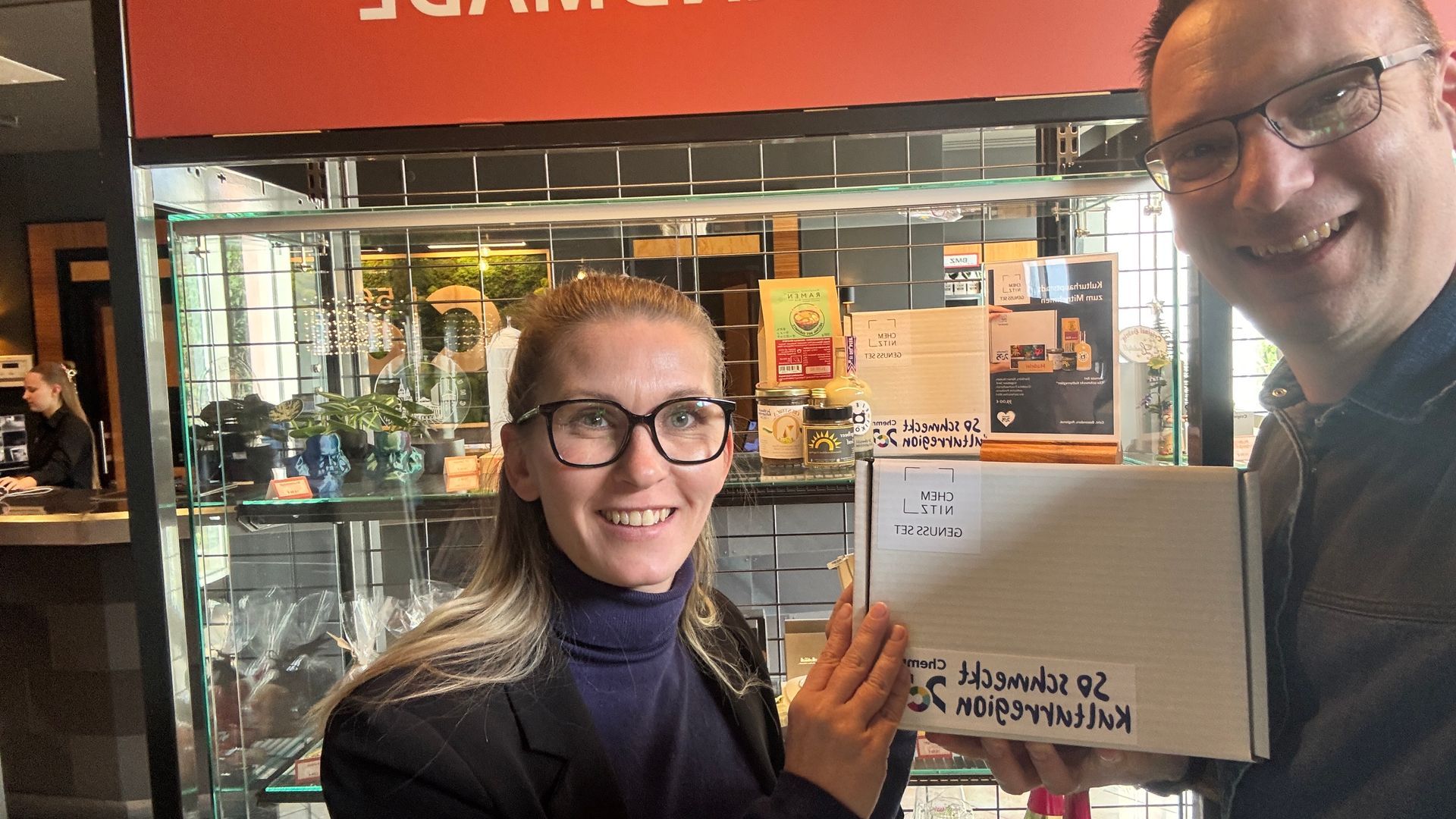 2 Personen, die vor einem Regal einen Kartn in der Hand halten.