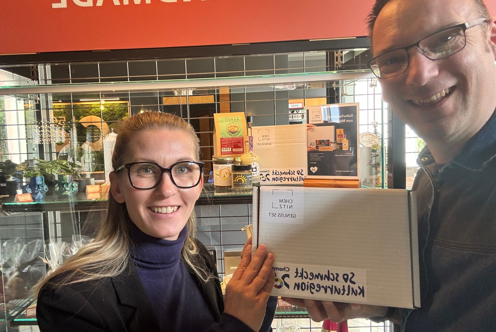 2 Personen, die vor einem Regal einen Kartn in der Hand halten.