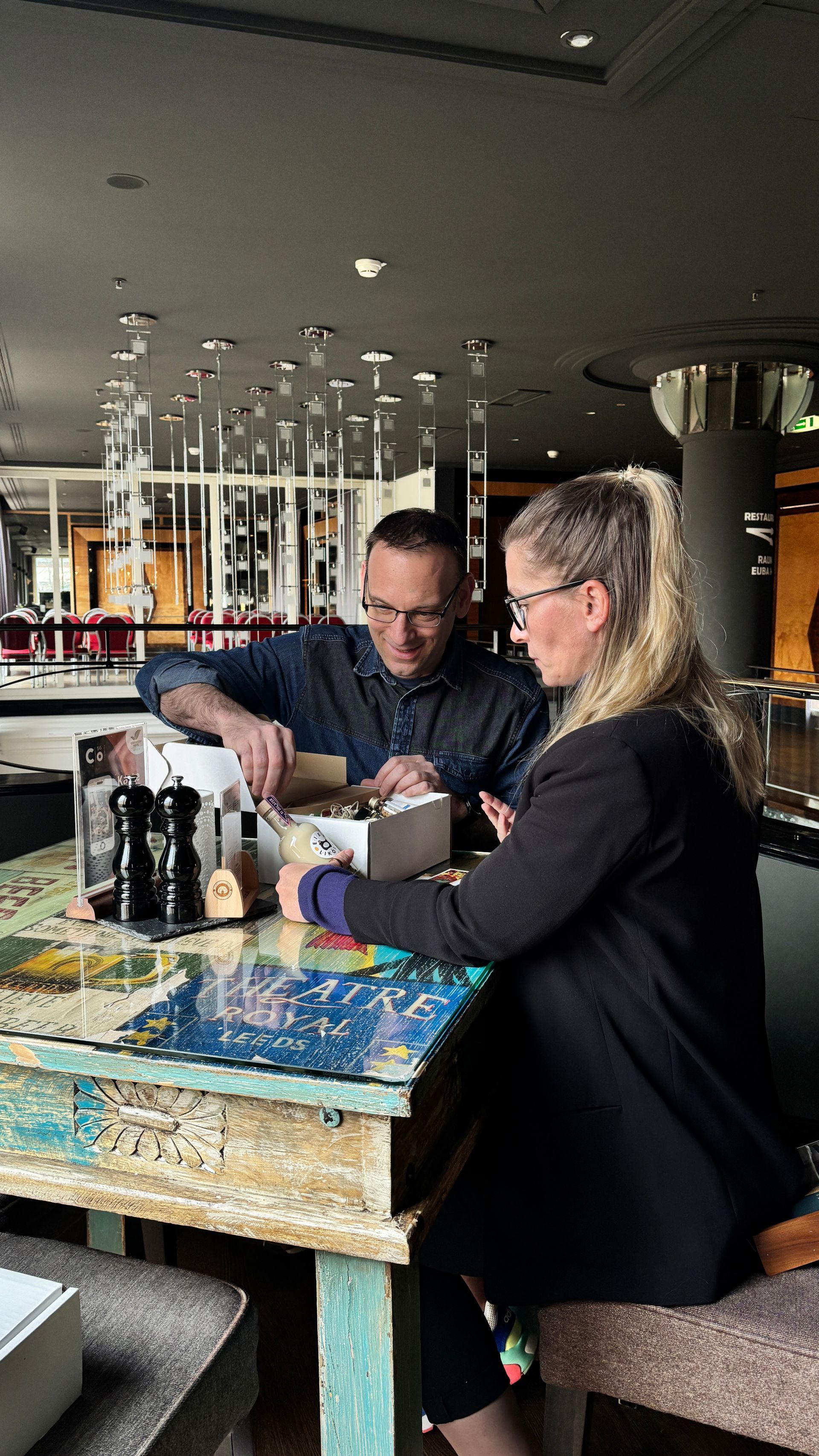 Eine Person in einem dunklen Blazer und eine andere in einem Jeanshemd arbeiten gemeinsam an einem strukturierten, farbenfrohen Tisch in einem schwach beleuchteten Raum.