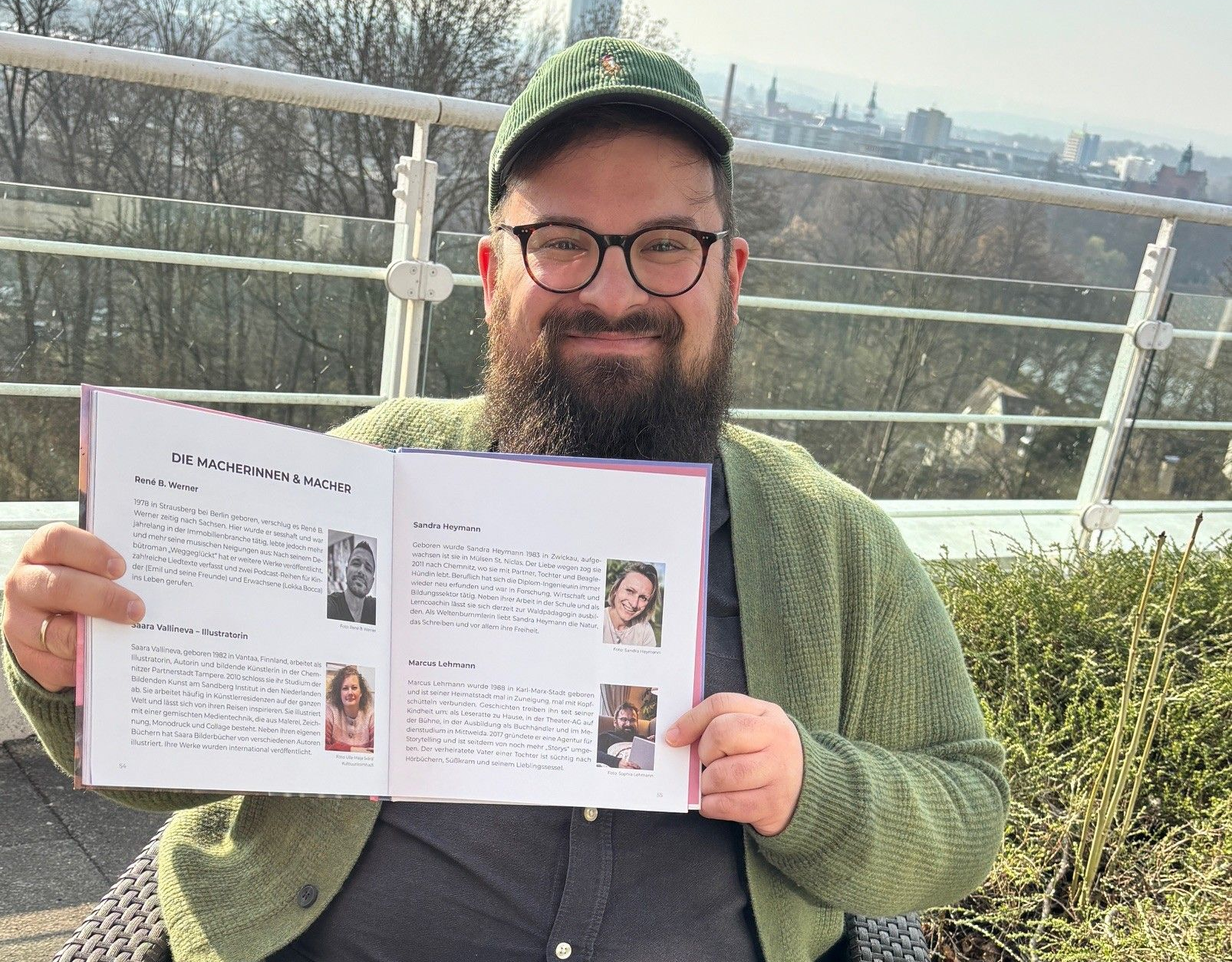 Ein Mann mit Bart und Brille hält im Freien vor der Kulisse der Skyline einer Stadt ein Buch mit Fotos & Texten.