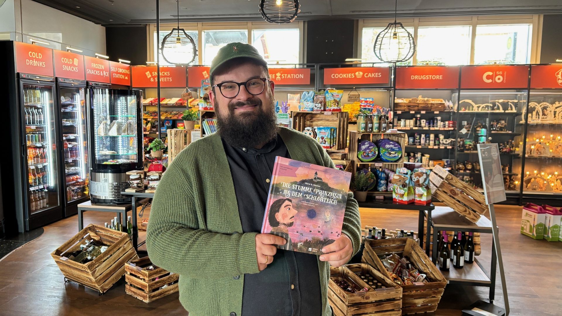 Eine Mann mit Bart und Brille, bekleidet mit einer grünen Strickjacke und Mütze, hält in einem Geschäft ein Buch in der Hand.