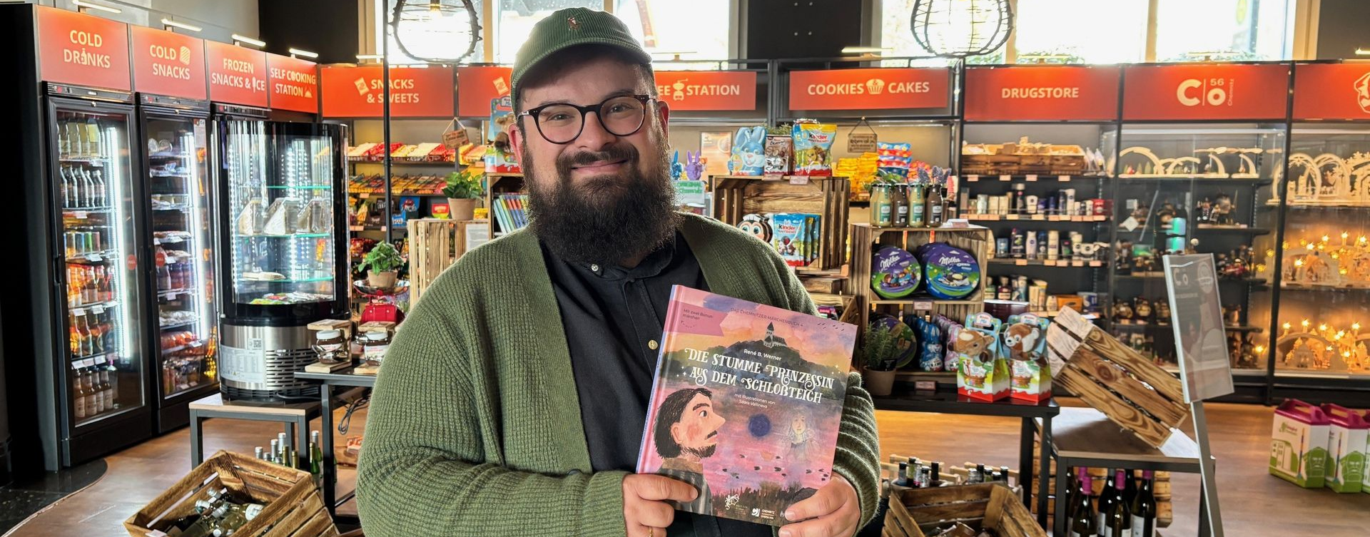 Ein Mann in grüner Strickjacke und Mütze hält in einem Geschäft, im Hintergrund sind Regale zu sehen, ein Buch hoch.
