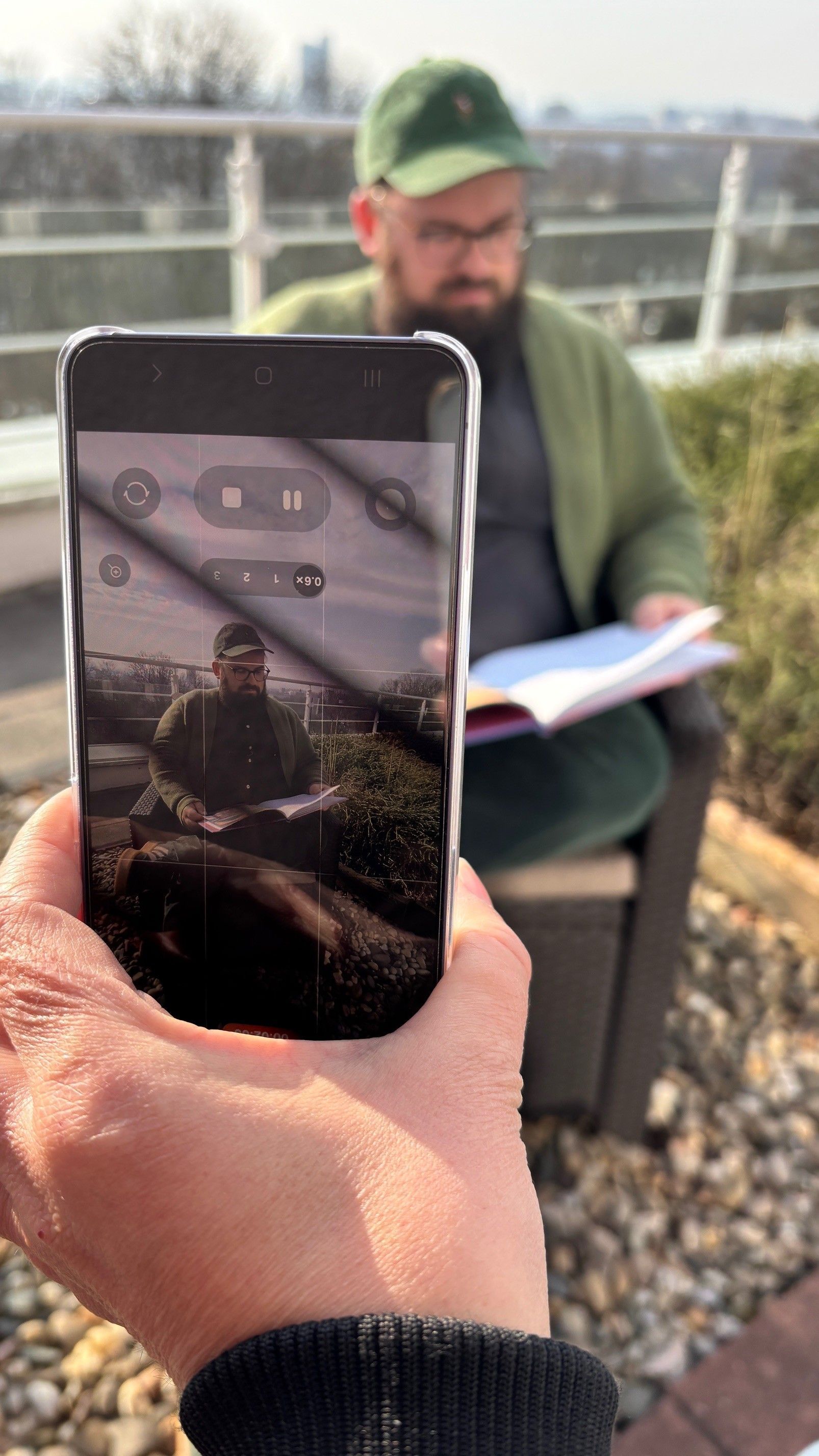 Jemand filmt mit ihrem Handy eine Person, die mit einem grünen Hut und Pullover bekleidet auf einer Terrasse sitzt.
