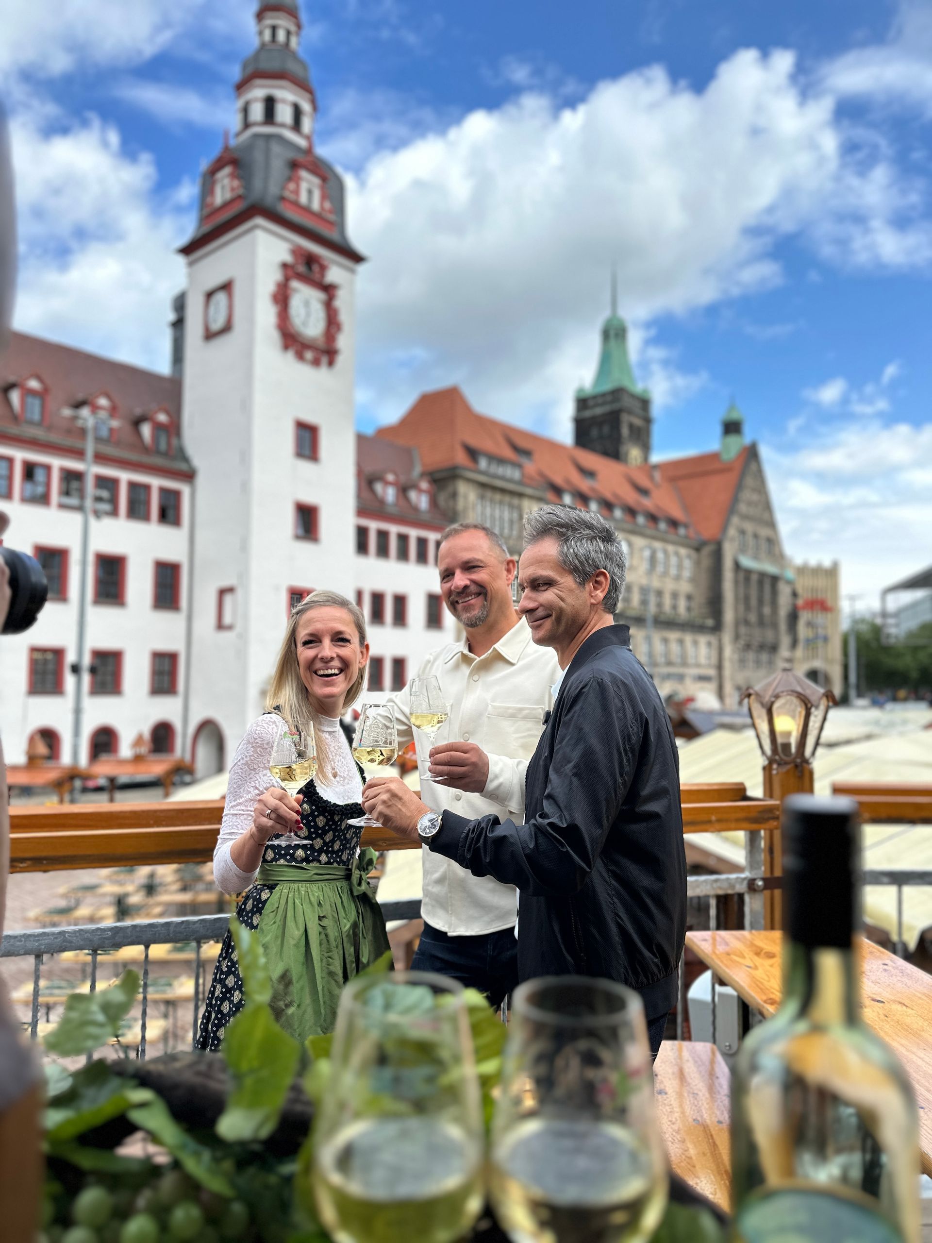 3 Menschen, die auf einer Terrasse mit historischen Gebäude und einem Glockenturm im Hintergrund ihre Weingläser erheben.