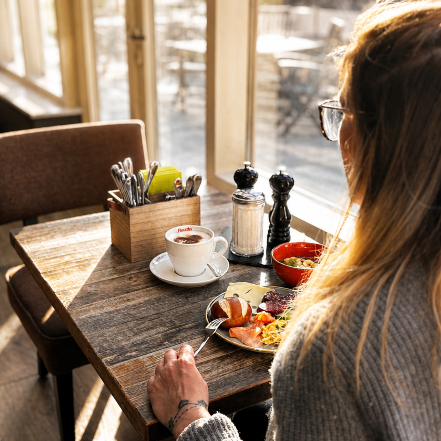 Person an einem Tisch mit Frühstück: Croissant, Eier, Speck, Obst, Tee.