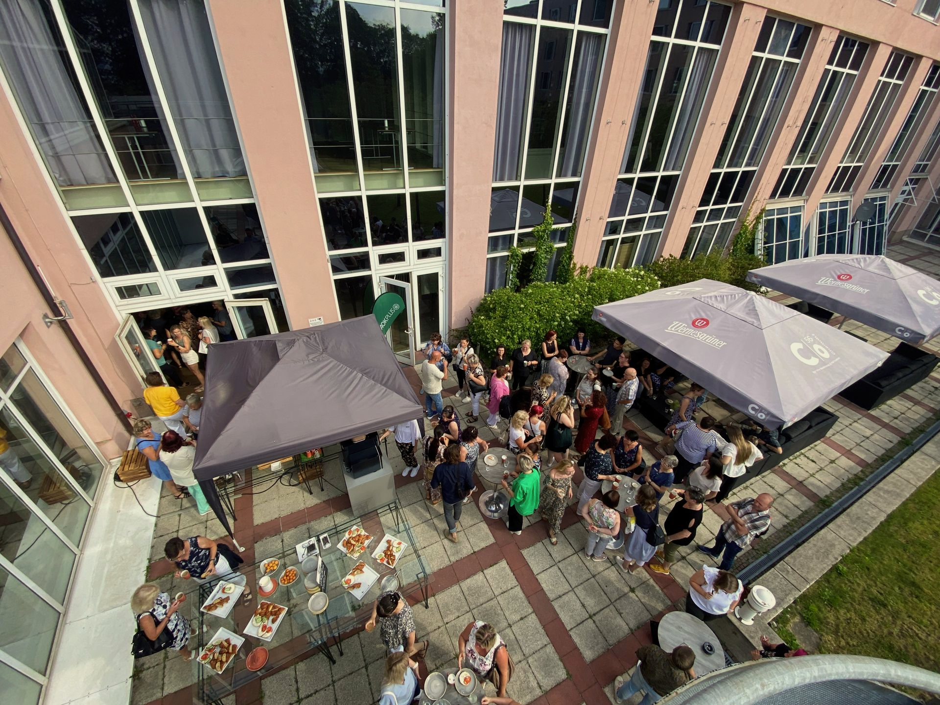 Eine gut besuchte Party im Innenhof neben einem modernen Gebäude, mit Tischen, Sonnenschirmen und vielen sitzenden Gästen.