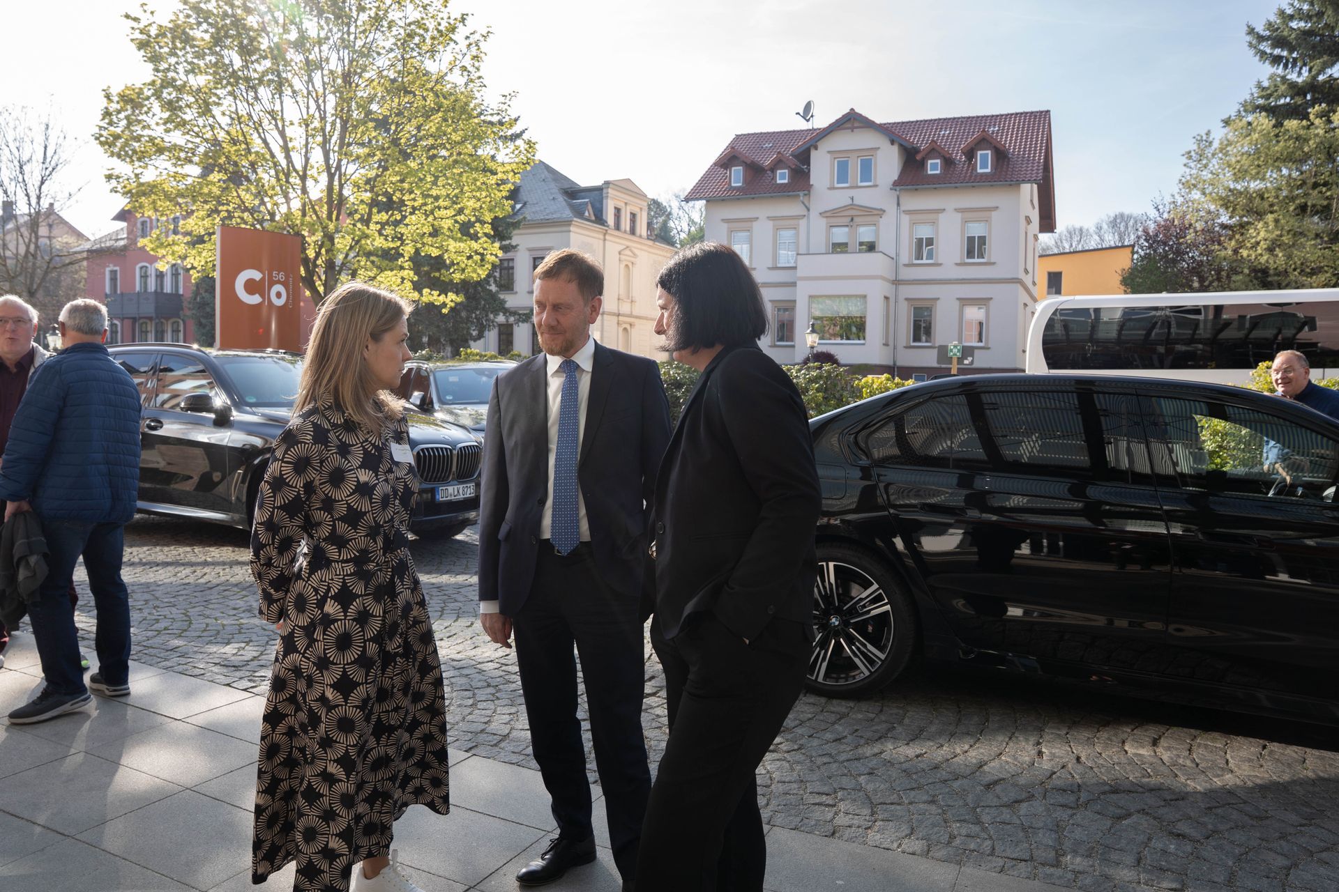 Drei Personen unterhalten sich neben einem schwarzen Auto auf einer sonnigen Straße, im Hintergrund 