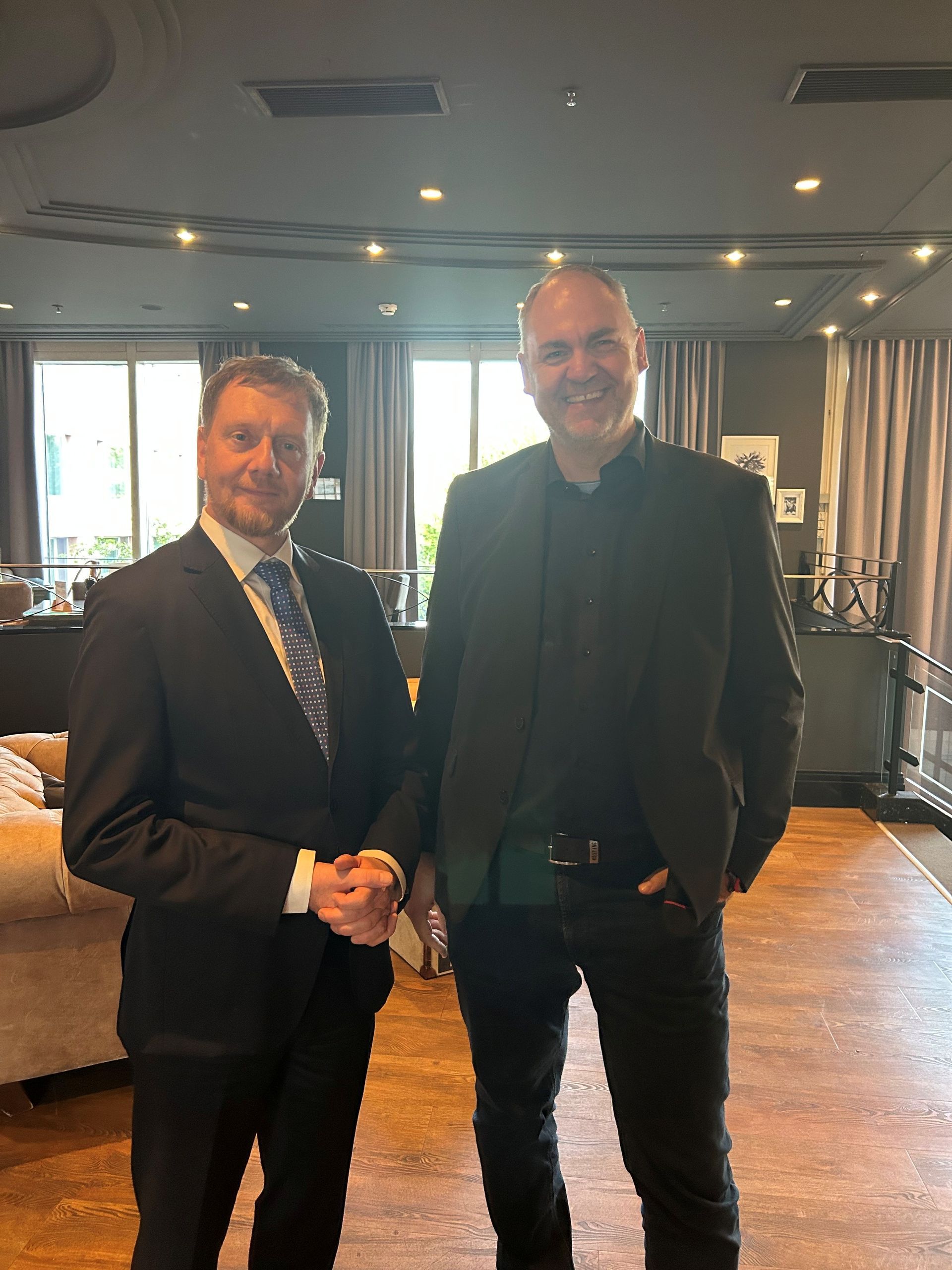 Zwei Männer in Anzügen stehen nebeneinander in einer warm beleuchteten Lounge, der eine lächelt, der andere hat die Hände in den Taschen.