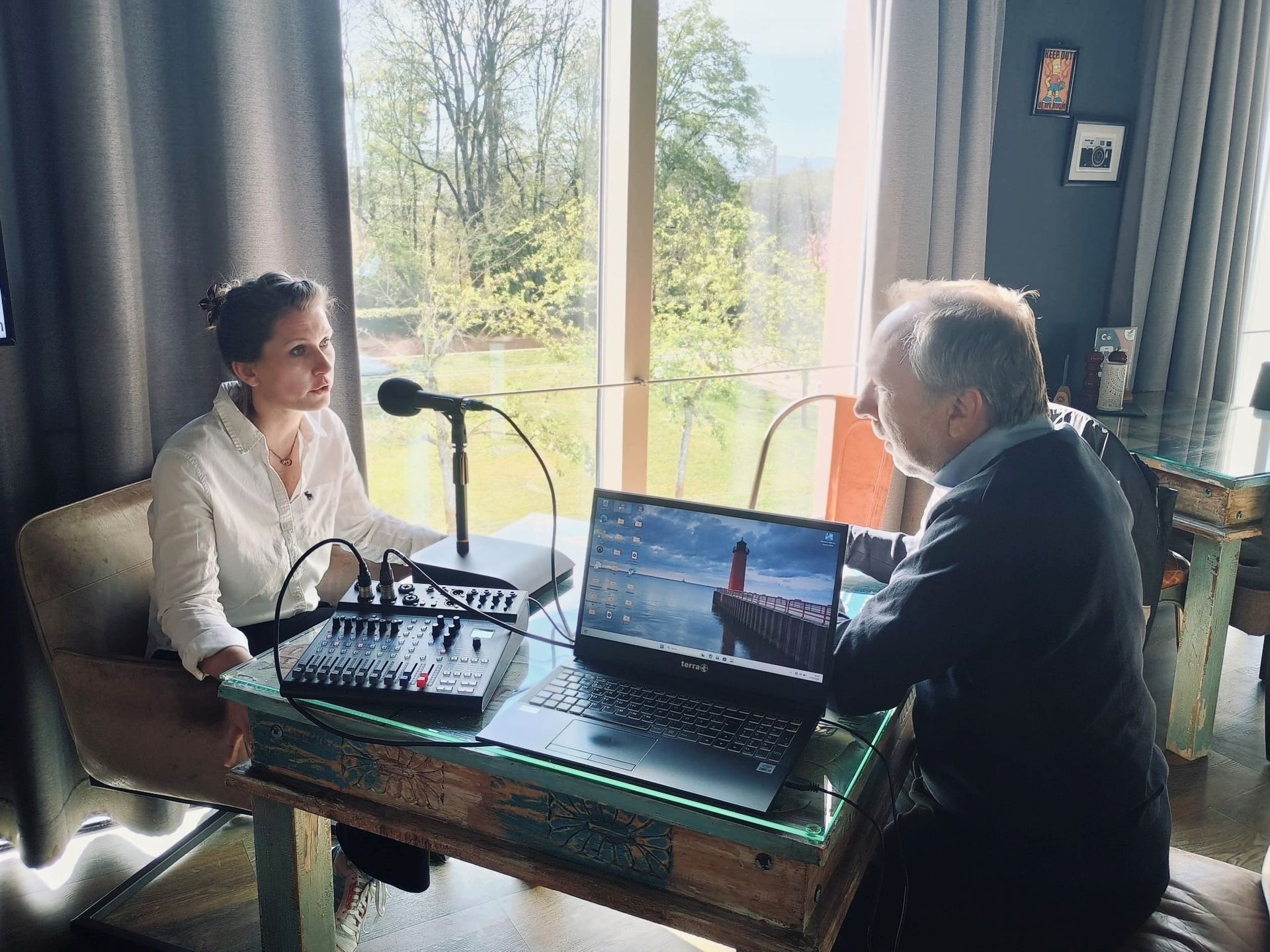 Zwei Personen sitzen an einem Tisch mit einem Laptop und Mikrofonen und unterhalten sich in einem he