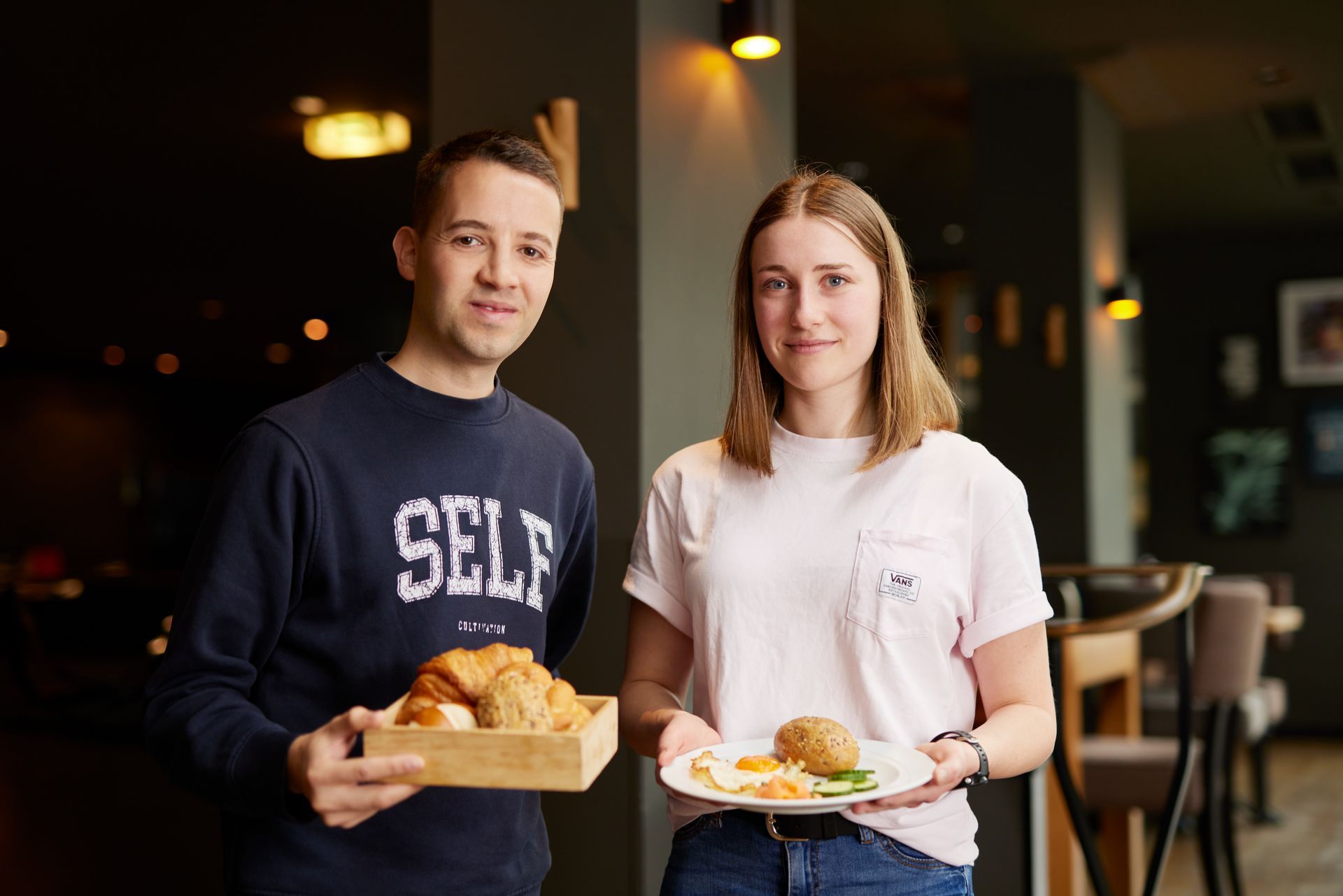 Zwei Café-Angestellte halten in einem schwach beleuchteten Restaurant eine Gebäckschachtel und ein belegtes Brötchen.
