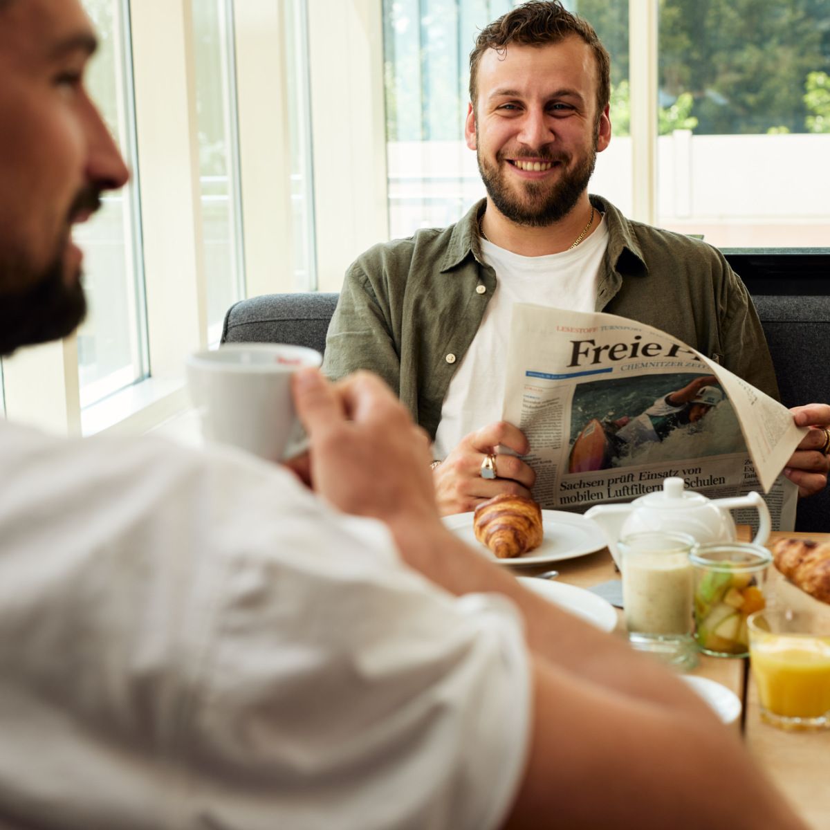 Zwei Männer genießen ihr Frühstück, einer lächelt beim Zeitunglesen, Croissants und Saft stehen auf dem Tisch.