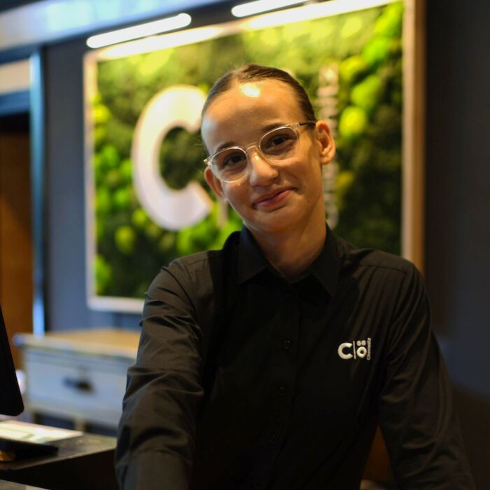 Eine Frau mit Brille und schwarzem Hemd lächelt hinter einem Empfangstresen; das „C“-Logo ist sichtbar.