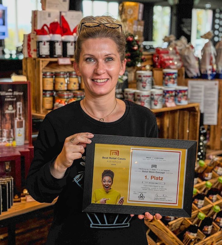 Frau mit gerahmtem Zertifikat, lächelnd in einem Geschäft, umgeben von Regalen mit Produkten.