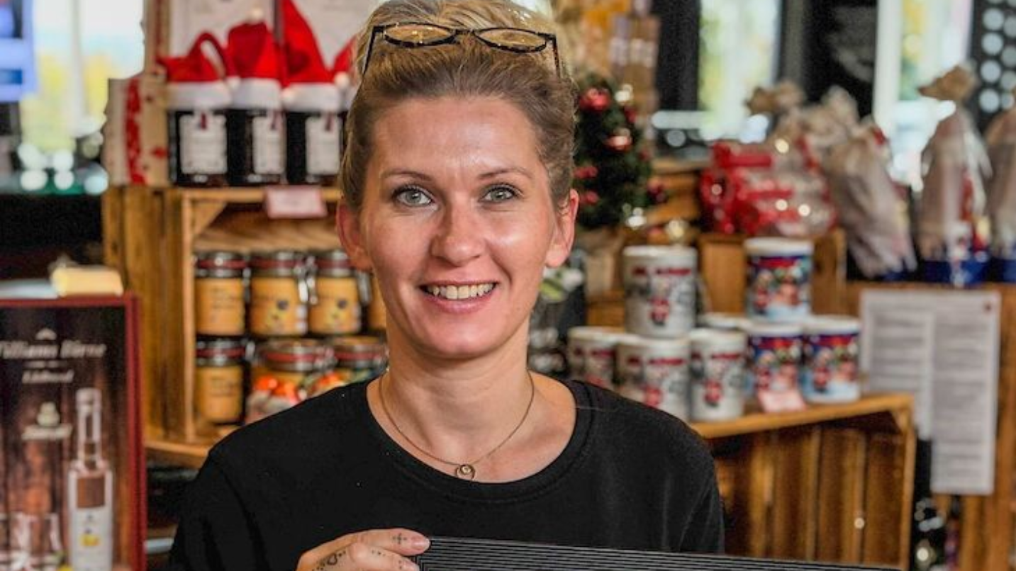 Eine Frau, die in einem Geschäft vor einem Regal mit Produkten lächelt, trägt eine Brille und ein schwarzes Hemd.