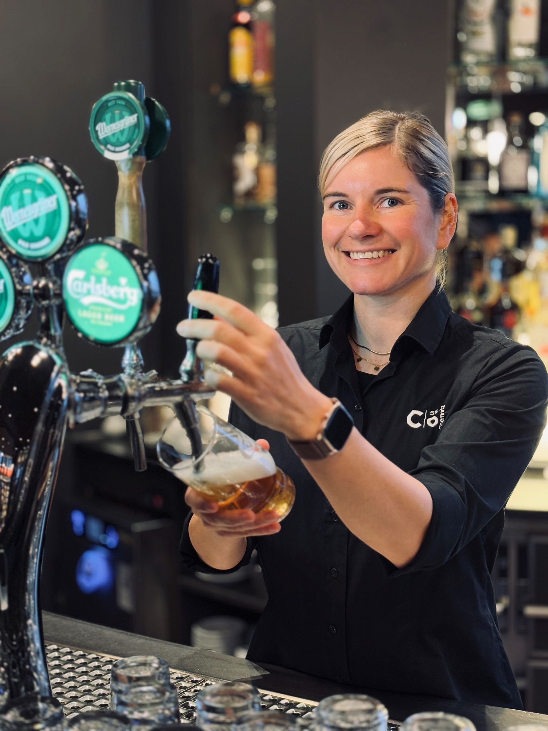 Eine Frau mit Brille und schwarzem Hemd lächelt hinter einem Empfangstresen; das „C“-Logo ist sichtbar.