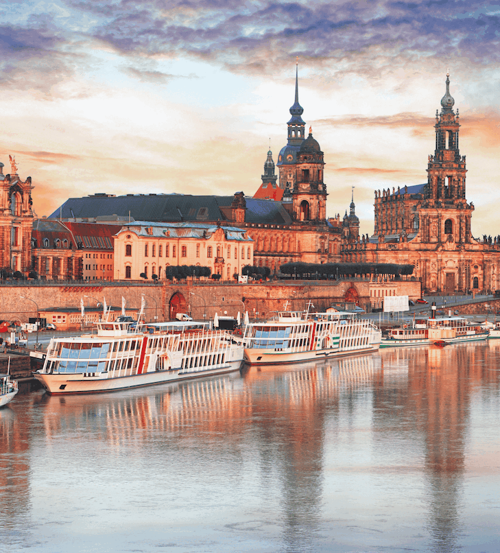 Stadtbild am Flussufer mit Booten und kunstvollen Gebäuden unter einem farbenprächtigen Himmel.