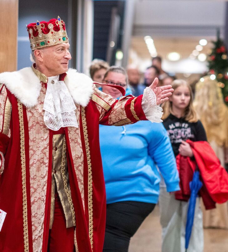 Ein Mann in reich verziertem rotem Gewand und mit Krone gestikulierend führt eine Menschenmenge in ein Gebäude.