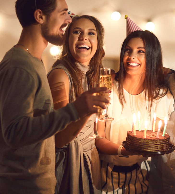 Drei Personen feiern: eine mit Kuchen und brennenden Kerzen, die anderen mit Getränken, alle lächelnd, Partystimmung.