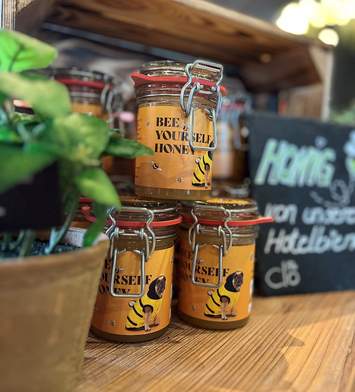 Honiggläser mit dem Etikett „Bee Yourself Honey“ und Metauf einem Holzregal. Pflanze im Vordergrund, Tafel im Hintergrund.