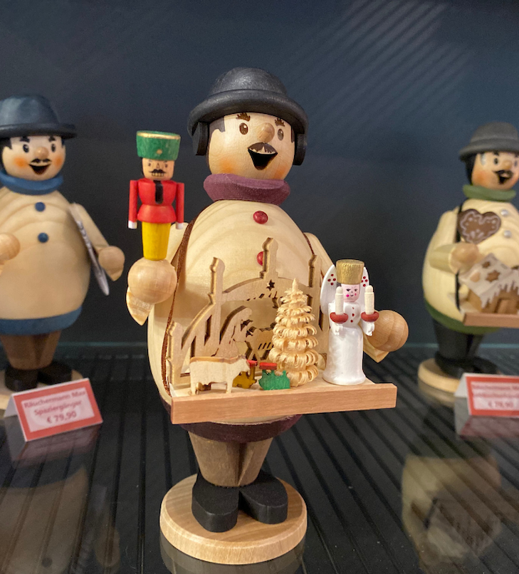 Eine Holzfigur hält ein Tablett mit Engel, Baum und Schaf. Im Hintergrund sind weitere Figuren zu sehen.