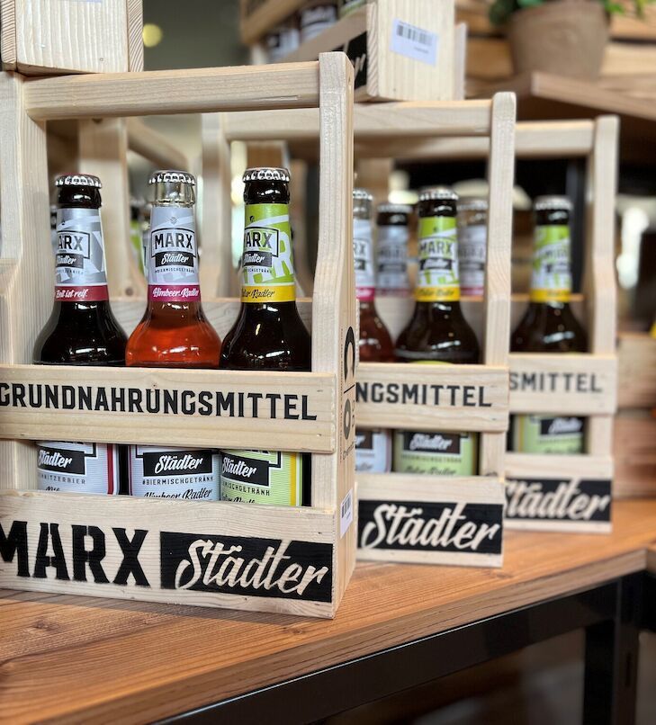 Holzkisten mit abgefüllten Getränken in einem Ladenregal, beschriftet mit „GRUNDAHRUNGSMITTEL“ und „MARX Städter“