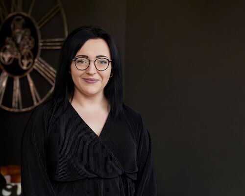 Eine Frau mit dunklen Haaren und Brille lächelt vor einer Uhr an einer dunklen Wand.