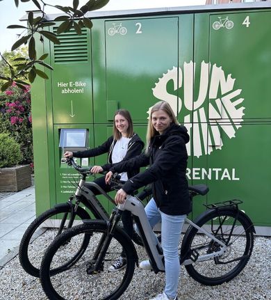 Zwei Frauen stehen mit E-Bikes vor einem grünen Fahrradverleihkiosk.