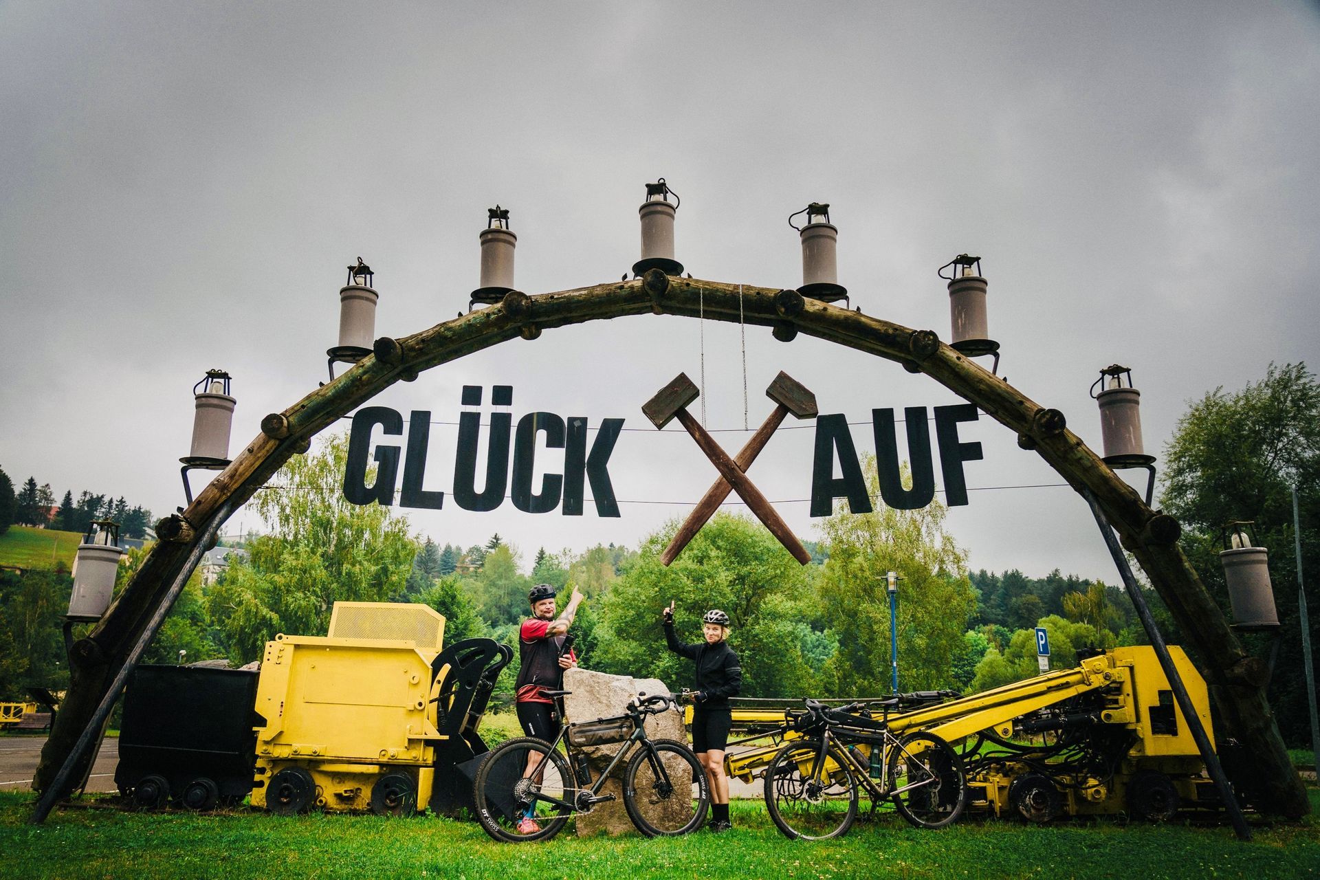 Zwei Radfahrer stehen unter einem Torbogen mit der Aufschrift „GLÜCK AUF“. Bergbauausrüstung ist zu sehen.