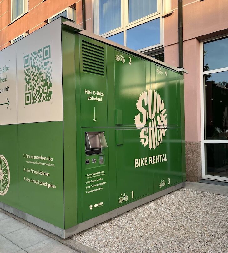 Grüne Fahrradverleihstation mit QR-Code und Text an der Gebäudeaußenwand.