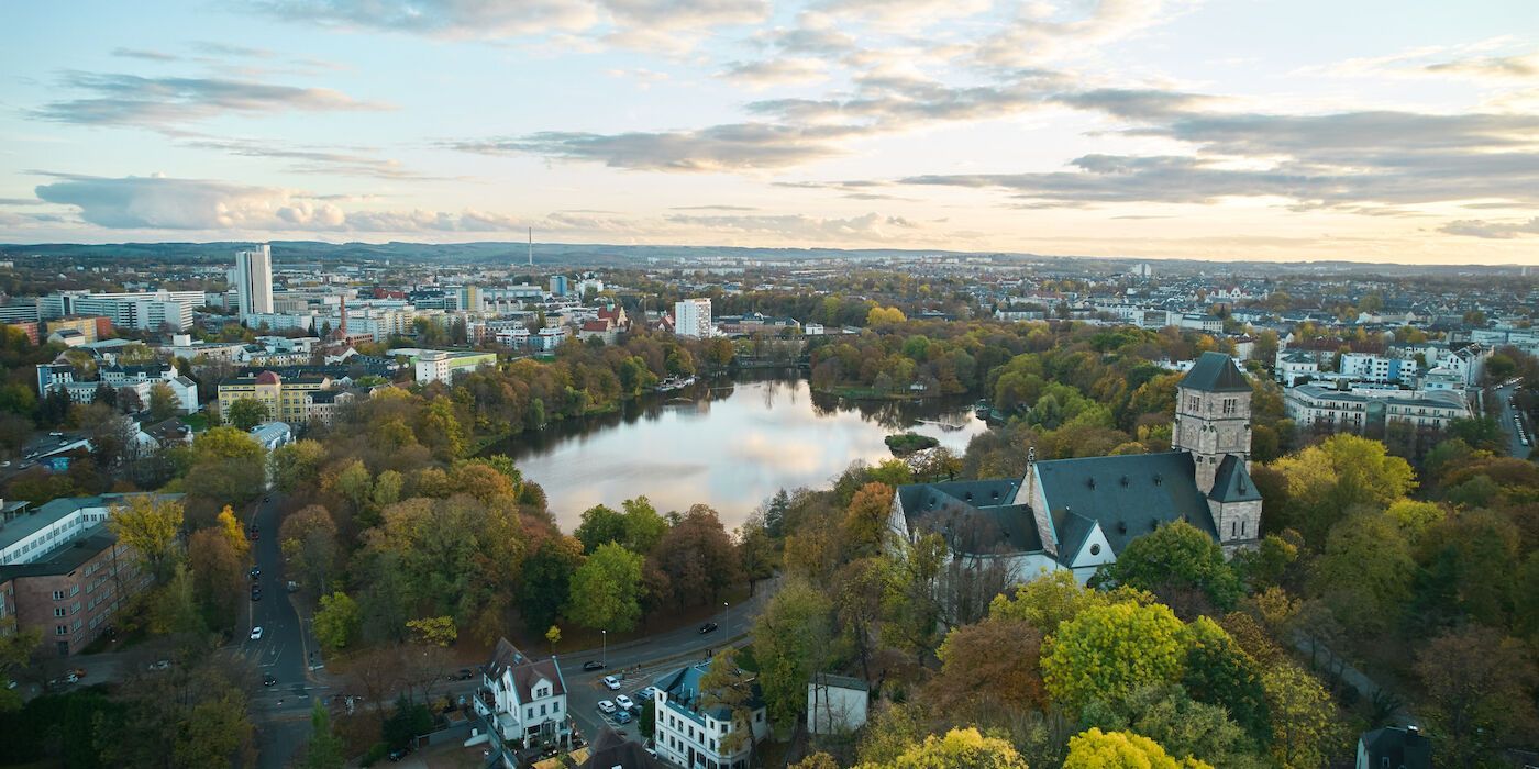 Luftaufnahme einer Stadt mit See, Bäumen und Gebäuden unter bewölktem Himmel.