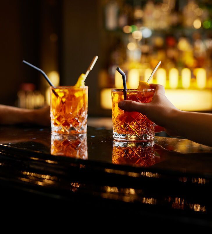 Zwei orangefarbene Cocktails mit Strohhalmen auf einer Bar, einer davon wird von einer Hand gehalten.