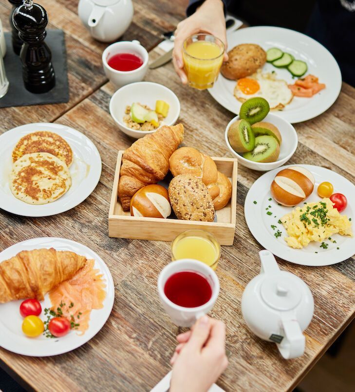 Auf einem Holztisch war ein Frühstücksbuffet angerichtet: Pfannkuchen, Eier, Bagels, Obst, Croissants, Saft und Tee.