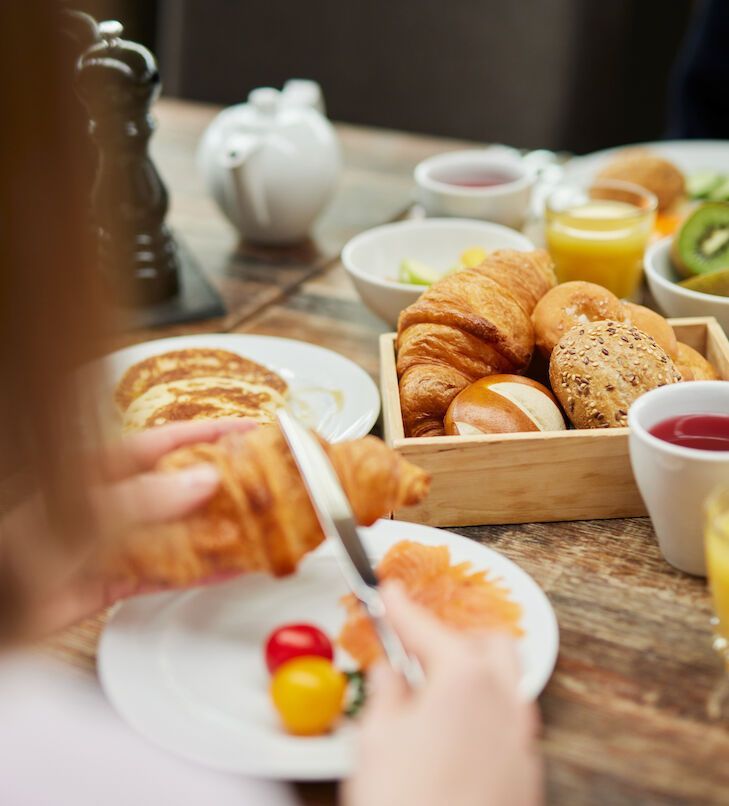 Eine Person schneidet an einem Frühstückstisch mit verschiedenen Speisen – Gebäck, Saft und Obst – ein Croissant.
