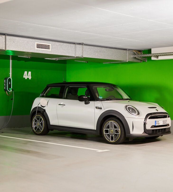 Ein weißer Mini Cooper EV lädt in einer Garage mit grünen Wänden. Ladestation an der Wand, Steckplatz Nummer 44.