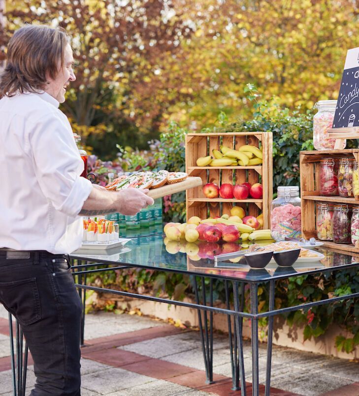 Eine Person stellt Essen auf einen Tisch; draußen gibt es eine Obstauslage und Snacks.