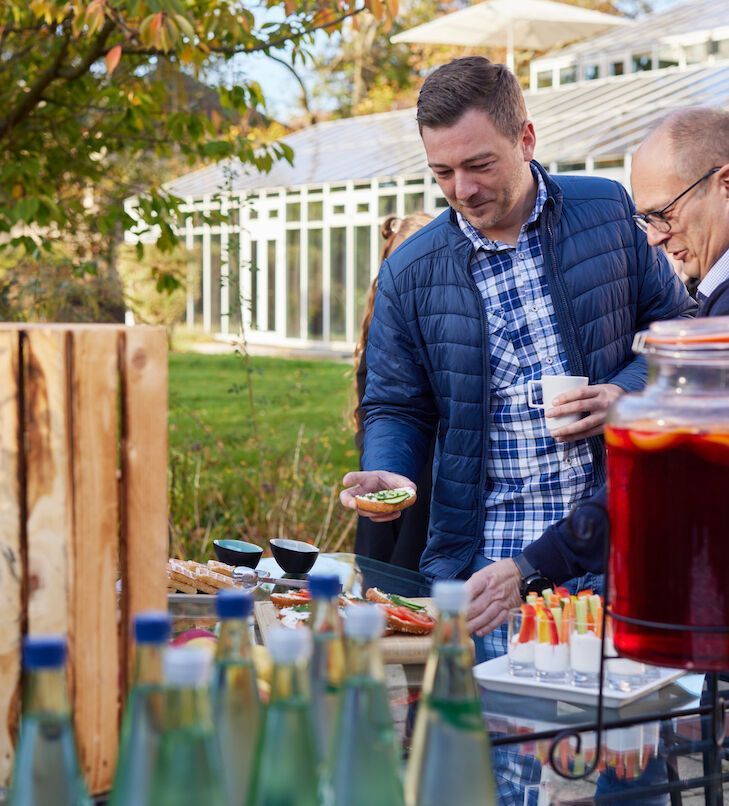Menschen bei einem Treffen im Freien mit Essen und Getränken. Ein Mann betrachtet das Essen. Im Hintergrund ein Glasgebäude.