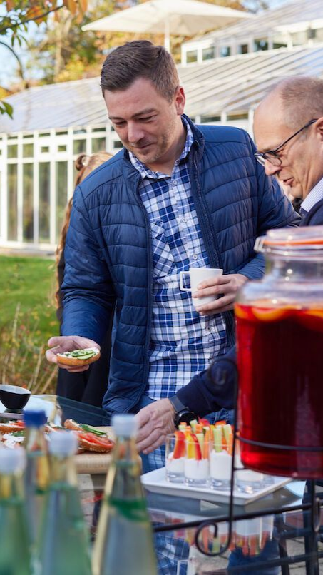 Männer an einem Buffet im Freien, die sich Speisen und Getränke aussuchen. Im Hintergrund ein Gewächshaus aus Glas und Bäume.