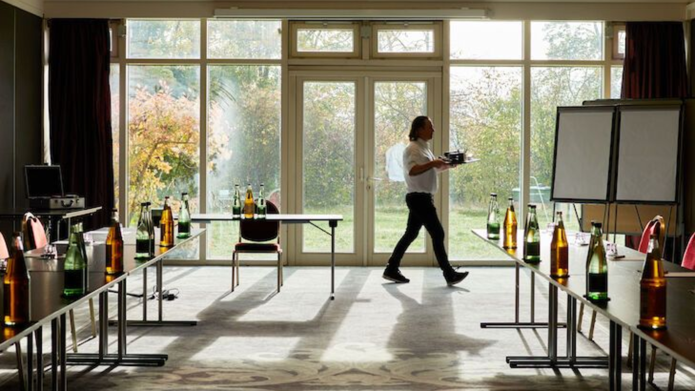 Ein Mann geht mit einem Tablett durch den Konferenzraum. Tische mit Flaschen und ein Flipchart sind zu sehen.