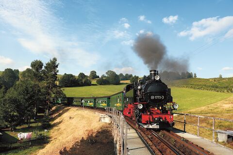 Eine Dampflok tuckert über die Gleise, durch grüne Hügel unter blauem Himmel, dunkler Rauch steigt auf.