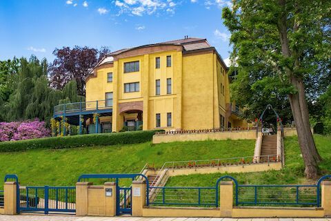 Gelbes Gebäude auf einem grasbewachsenen Hügel mit blauem Zaun. Bäume und blauer Himmel im Hintergrund.