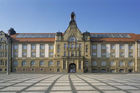 Großes beiges Gebäude mit vielen Fenstern, gewölbtem Eingang und klarem blauen Himmel.