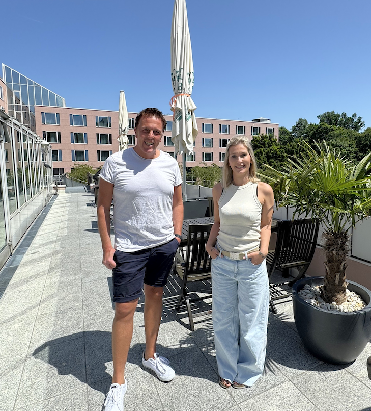 Zwei Personen stehen an einem sonnigen Tag draußen auf einer Terrasse. Der Mann trägt Shorts, die Frau Jeans.