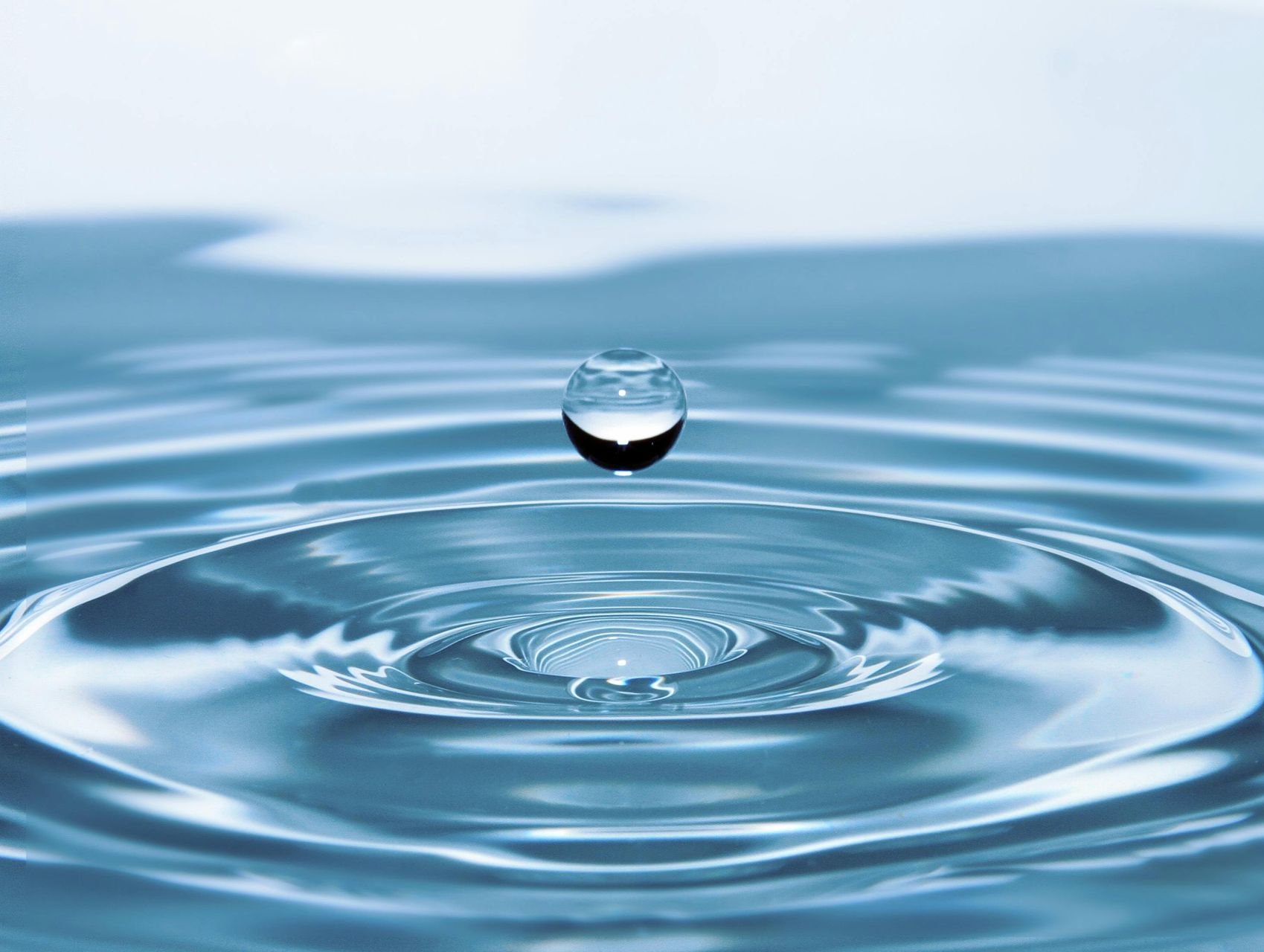 Ein Wassertropfen spritzt in ruhiges blaues Wasser und erzeugt konzentrische Wellen.
