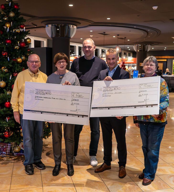 Menschen halten große Schecks in der Nähe eines geschmückten Weihnachtsbaums in einem Innenraum.